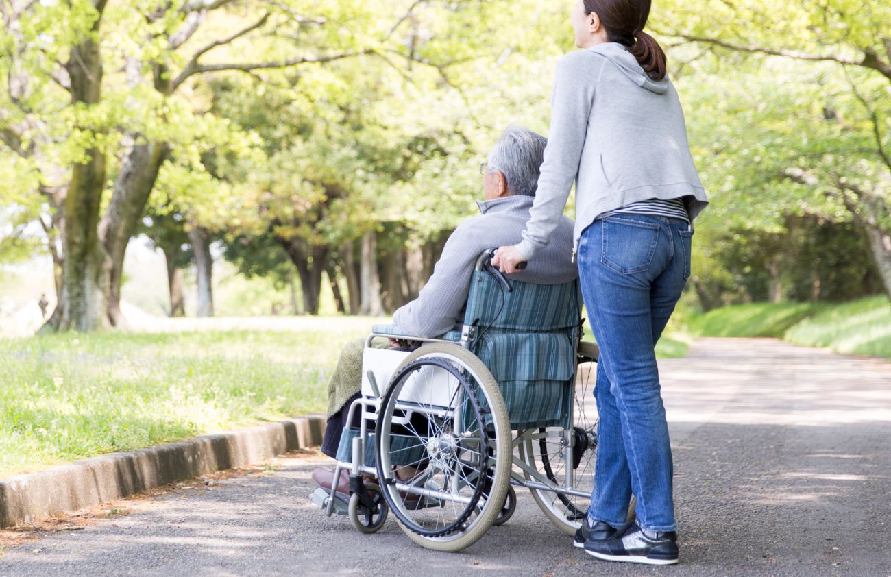 Eine Frau schiebt einen älteren Mann in einem Rollstuhl, während sie gemeinsam im Freien unterwegs sind.