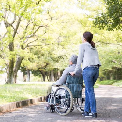 Eine Frau schiebt einen älteren Mann im Rollstuhl einen Weg entlang, umgeben von einer ruhigen, natürlichen Umgebung.
